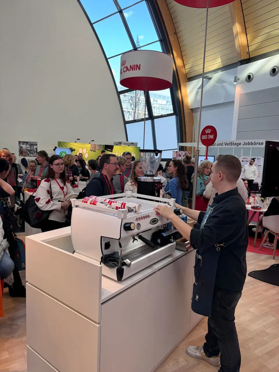 Messestand mit Coffee Bar, ein Barista bedient Gäste am Counter, Besucher im Gespräch davor