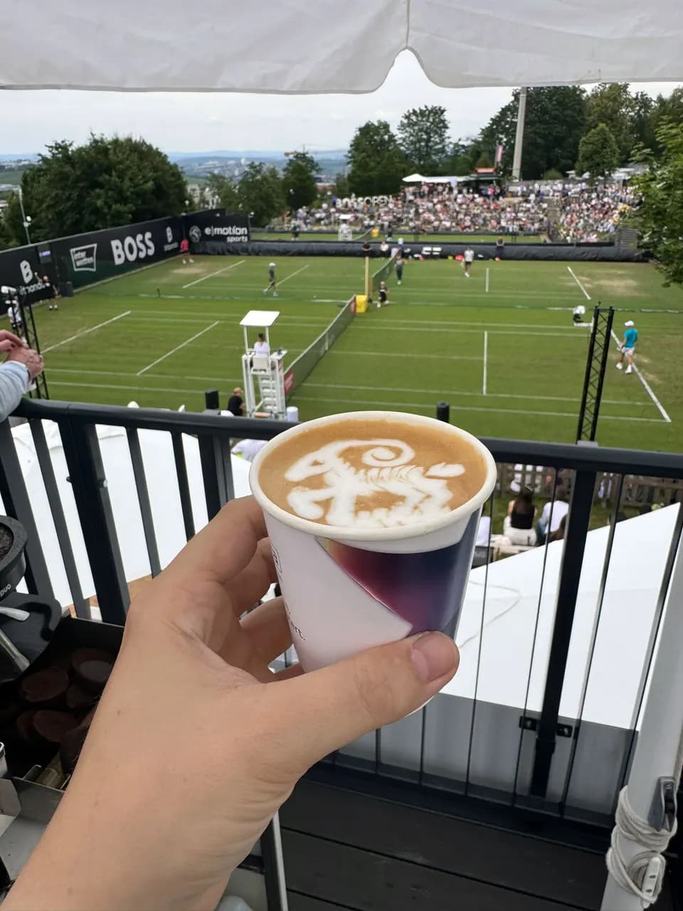 Hand hält einen Becher mit Latte Art über der Tribüne eines Sportevents