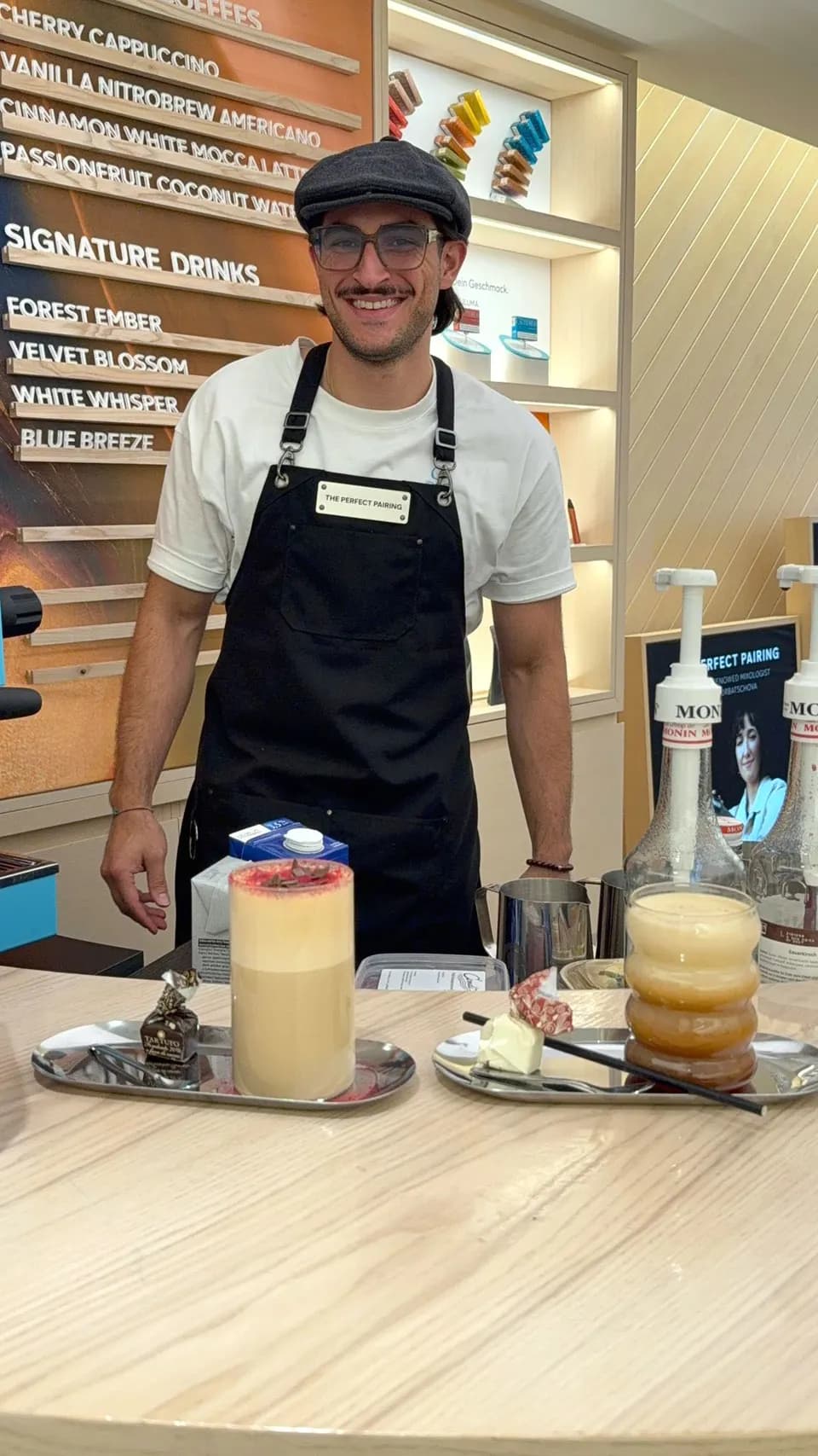 Barista hinter einer Theke mit Sirupflaschen und einer Getränketafel